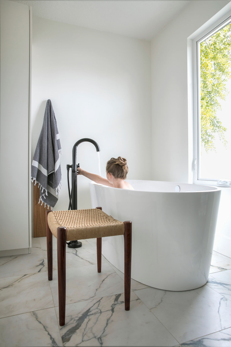 Baño minimalista con bañera exenta blanca, grifería negra, ventanal de luz natural y piso de mármol – FRANCO HomeServices